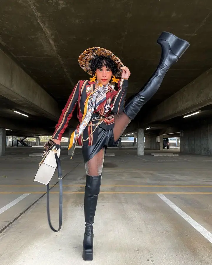 A person with curly hair and a wide-brimmed hat poses in a parking garage, wearing a colorful striped jacket, a patterned…