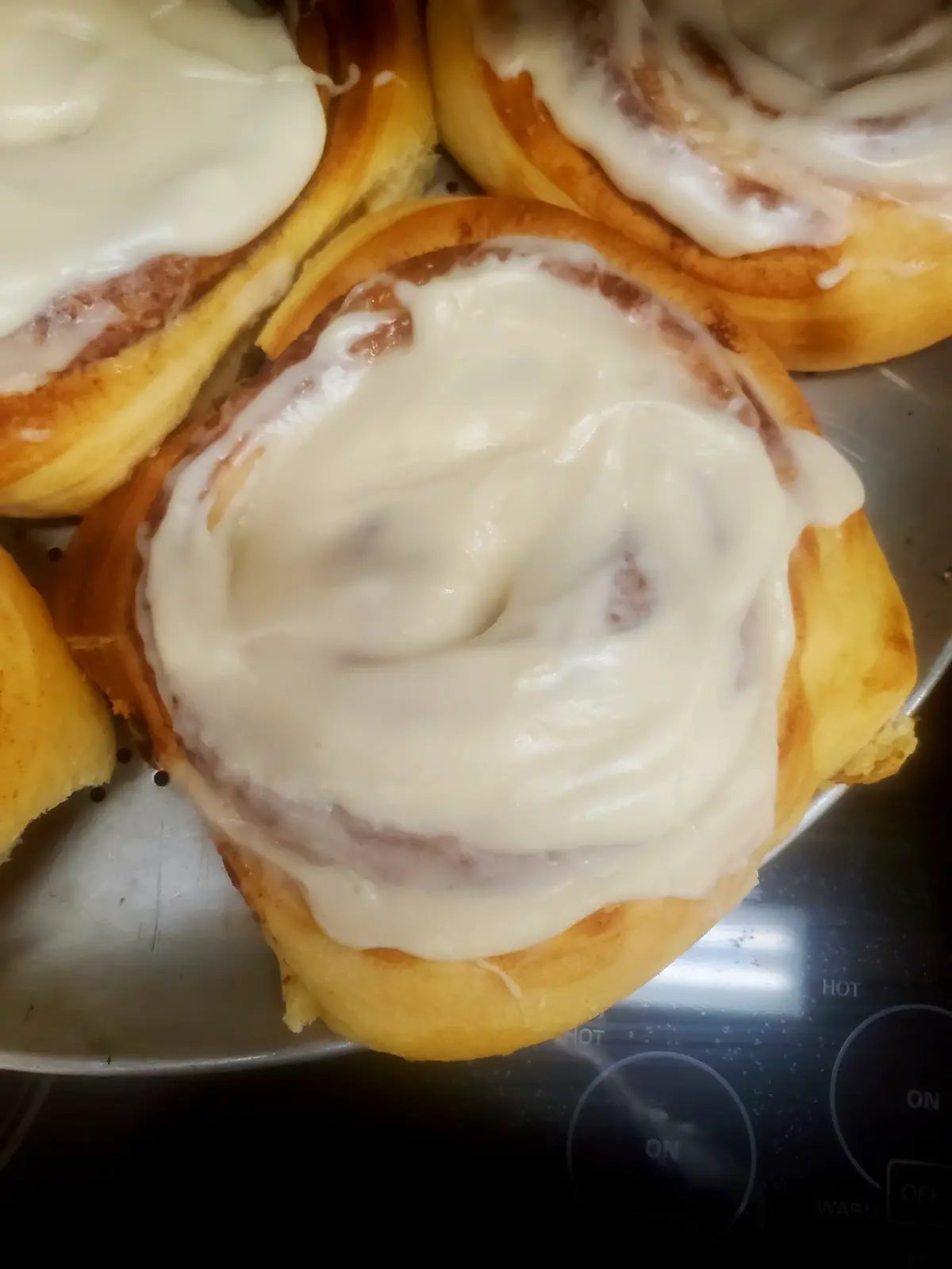 A close-up photo shows several freshly baked cinnamon rolls with white icing on a metal baking sheet, resting on a black…