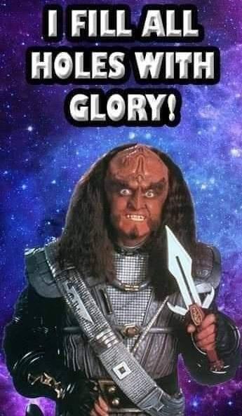 A menacing, bald, orange-skinned alien with long dark hair and a fierce expression, wearing ornate armor and holding a…