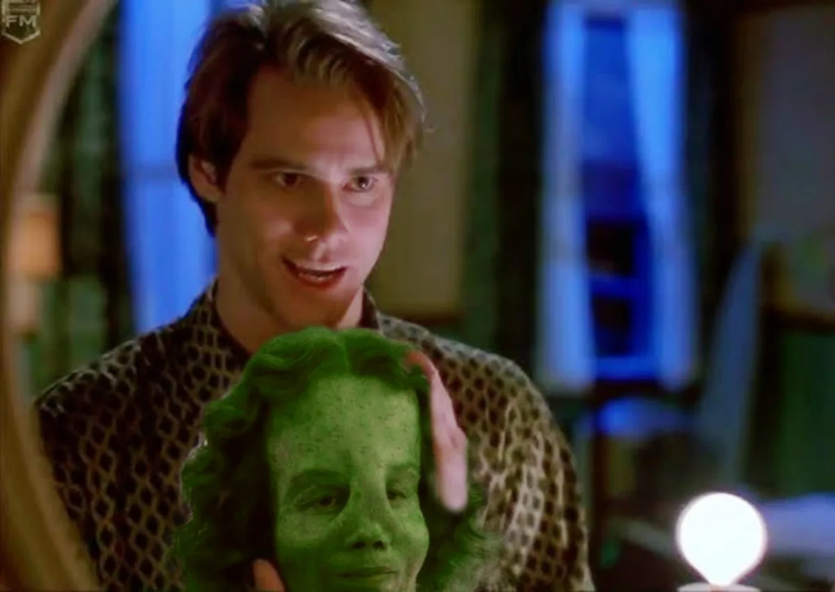 A man in a patterned shirt holds a green-painted human face mask, looking at it with a slight smile. The overlaid text reads…