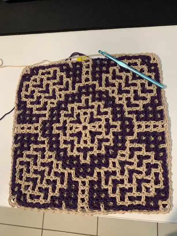 A square piece of crocheted fabric with a geometric, repeating pattern in purple and beige yarn, resembling a QR code, lies…