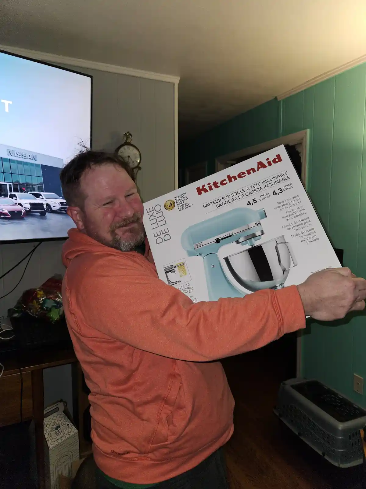 A man in an orange hoodie holds up a large KitchenAid stand mixer box, smiling proudly. The box features a light blue mixer…