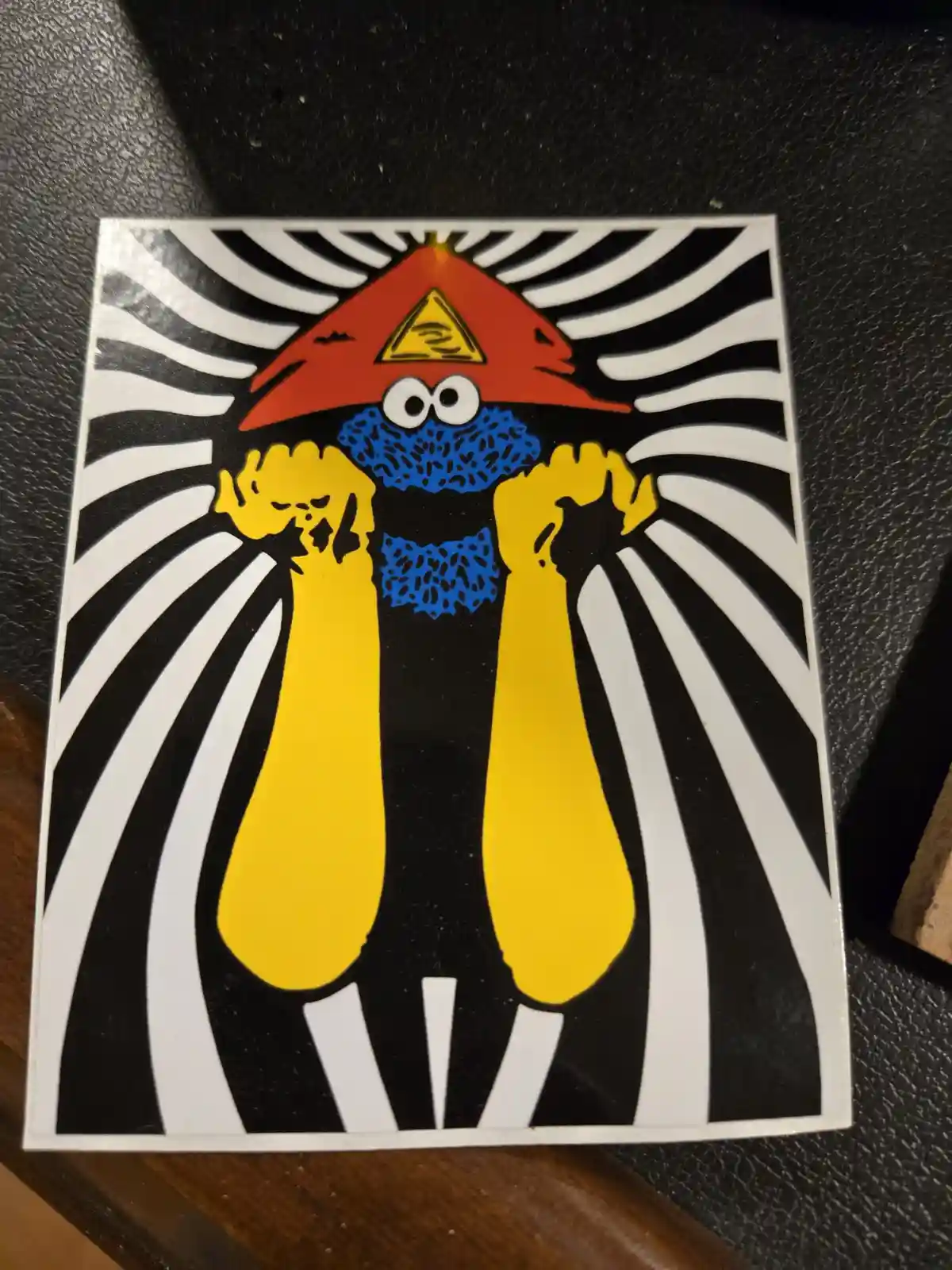 A sticker depicts a cartoon character with a red pointed hat featuring a yellow triangle with an eye, a blue beard, and…