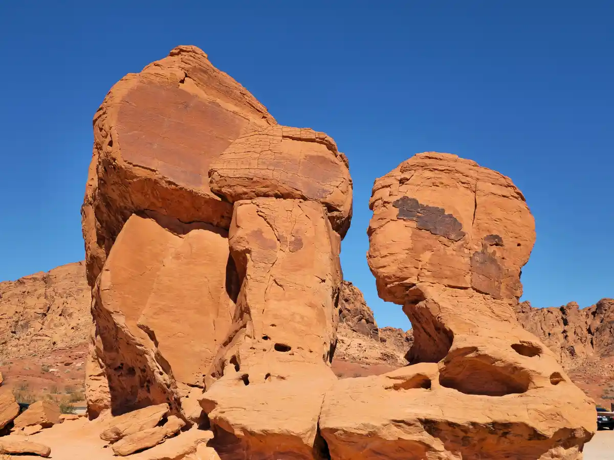 Two large, orange-red sandstone rock formations in a desert under a clear blue sky, shaped to resemble a man and a woman in…