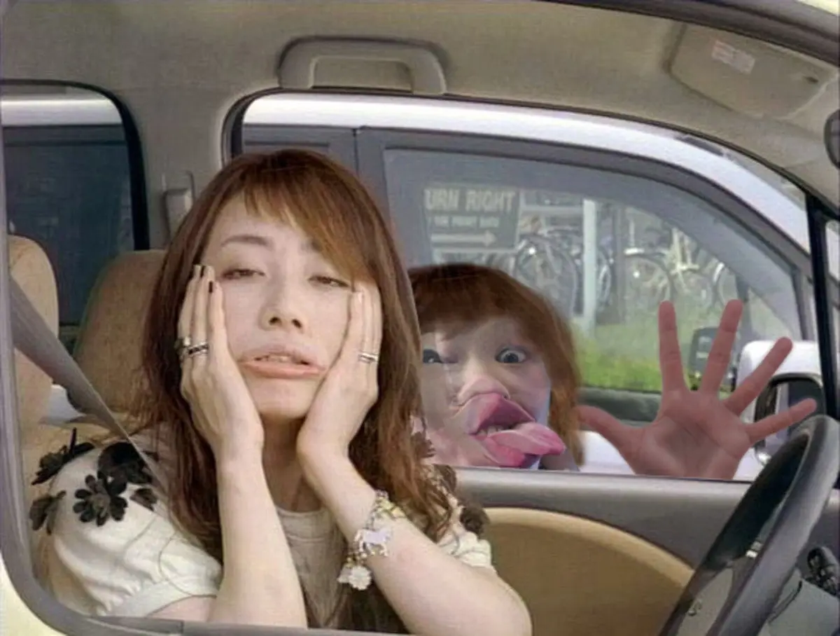 A woman sits in a car, hands on her face looking exhausted or annoyed, while a distorted, cartoonish version of her face…