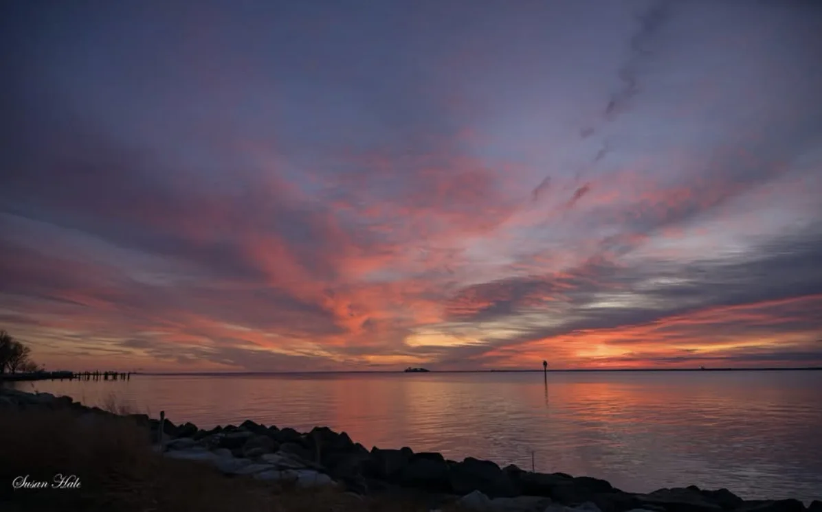 A serene sunset over water with vibrant pink, orange, and purple clouds reflecting on calm water, framed by a rocky…