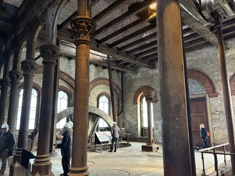 The image shows the interior of a large, historic industrial building with massive, ornate, rusted iron columns and arched…