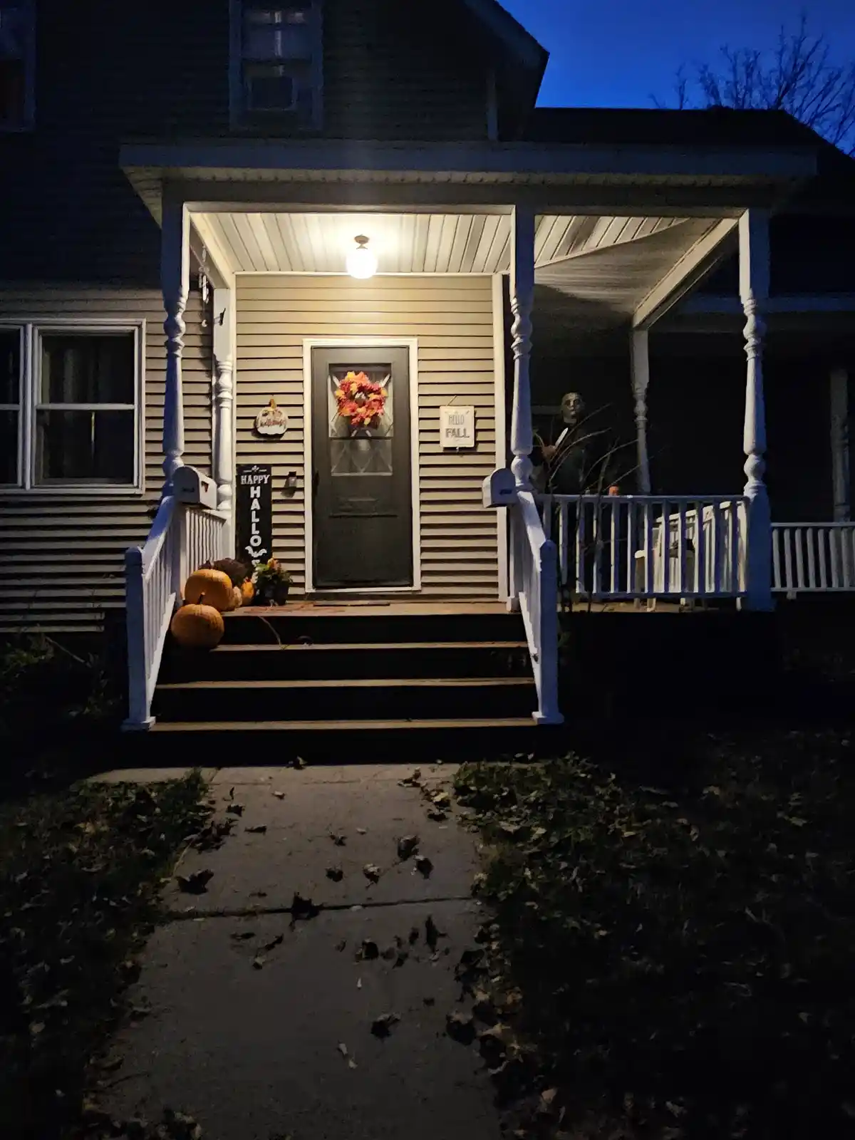 A house porch at dusk decorated for fall with pumpkins, a wreath, and a 'Happy Halloween' sign, but a Michael Myers…
