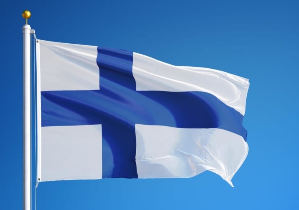 a clear blue sky with the national flag of finland waving on a white flagpole, featuring a blue cross on a white background…