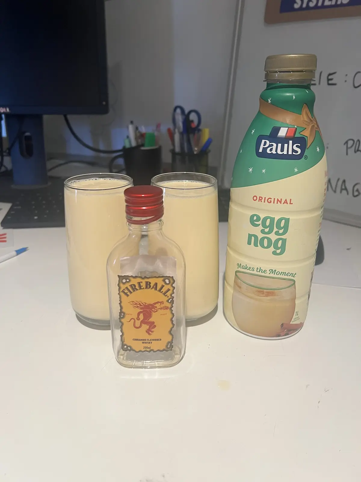A desk setup shows two glasses of creamy egg nog, a bottle of Pauls Original egg nog, and a small bottle of Fireball…