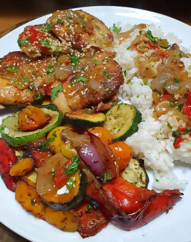 A close-up photo of a white plate filled with a colorful, glossy meal featuring glazed chicken pieces topped with sesame…