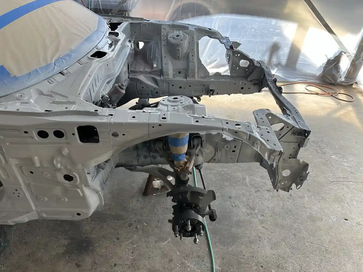 A car's metal body frame is shown stripped down to bare metal and freshly painted gray, resting on a concrete floor in a…