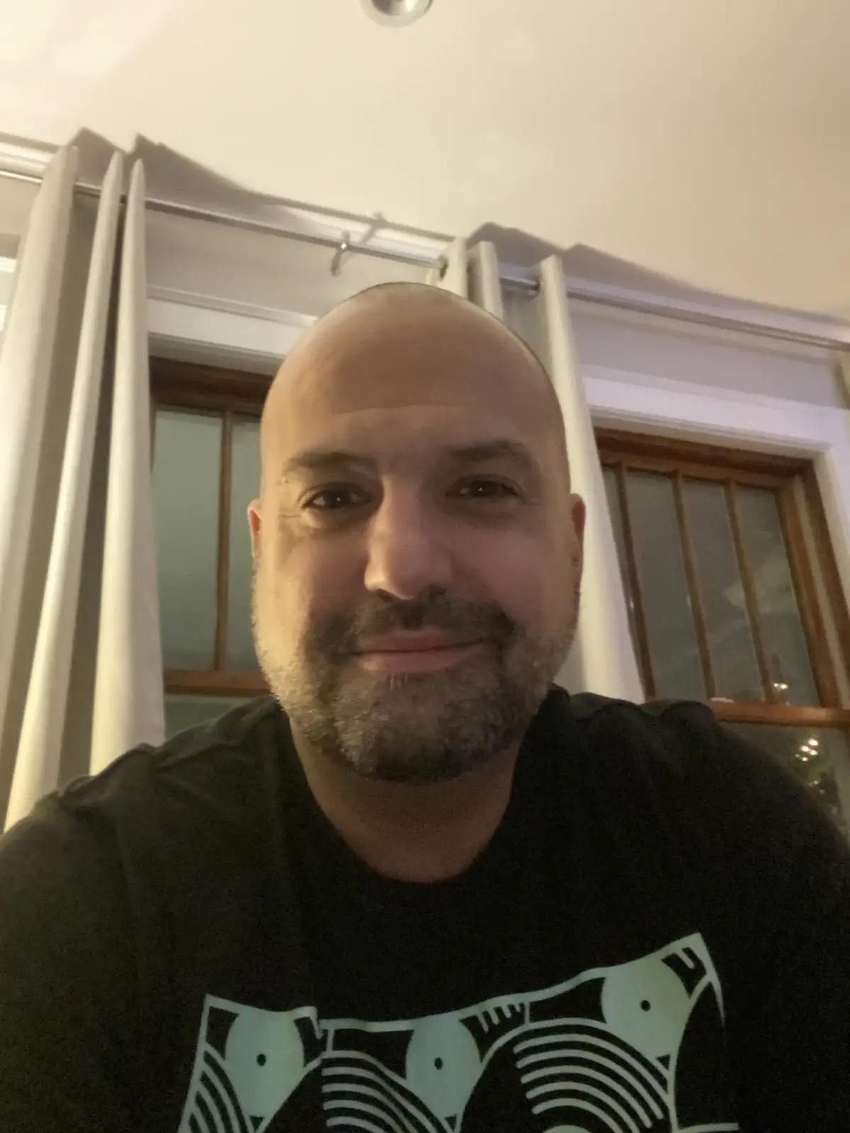 A bald man with a short beard smiles at the camera while wearing a black t-shirt with a light-colored graphic. Behind him…