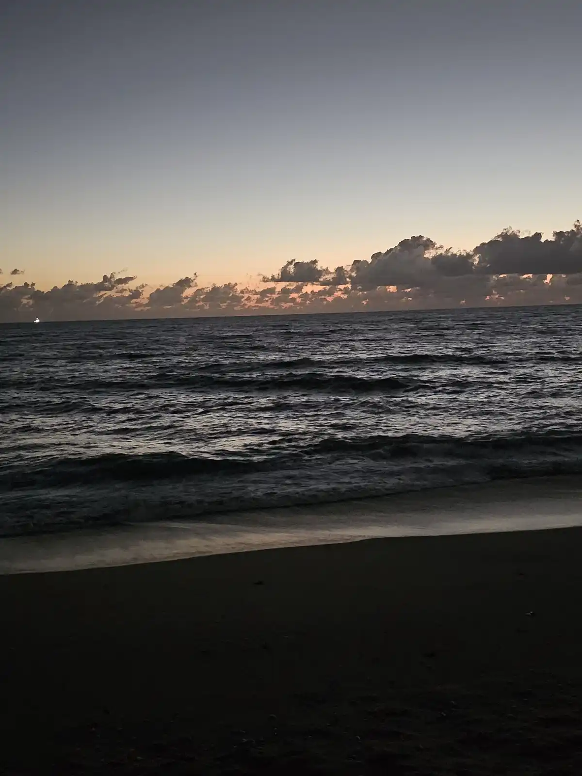 A serene beach sunset photo with dark water, a sandy shore, and clouds tinged with orange near the horizon. A small boat's…