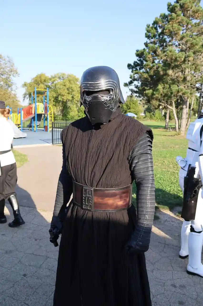 A person in a full Kylo Ren costume from Star Wars stands in a park, with a playground visible in the background and a…