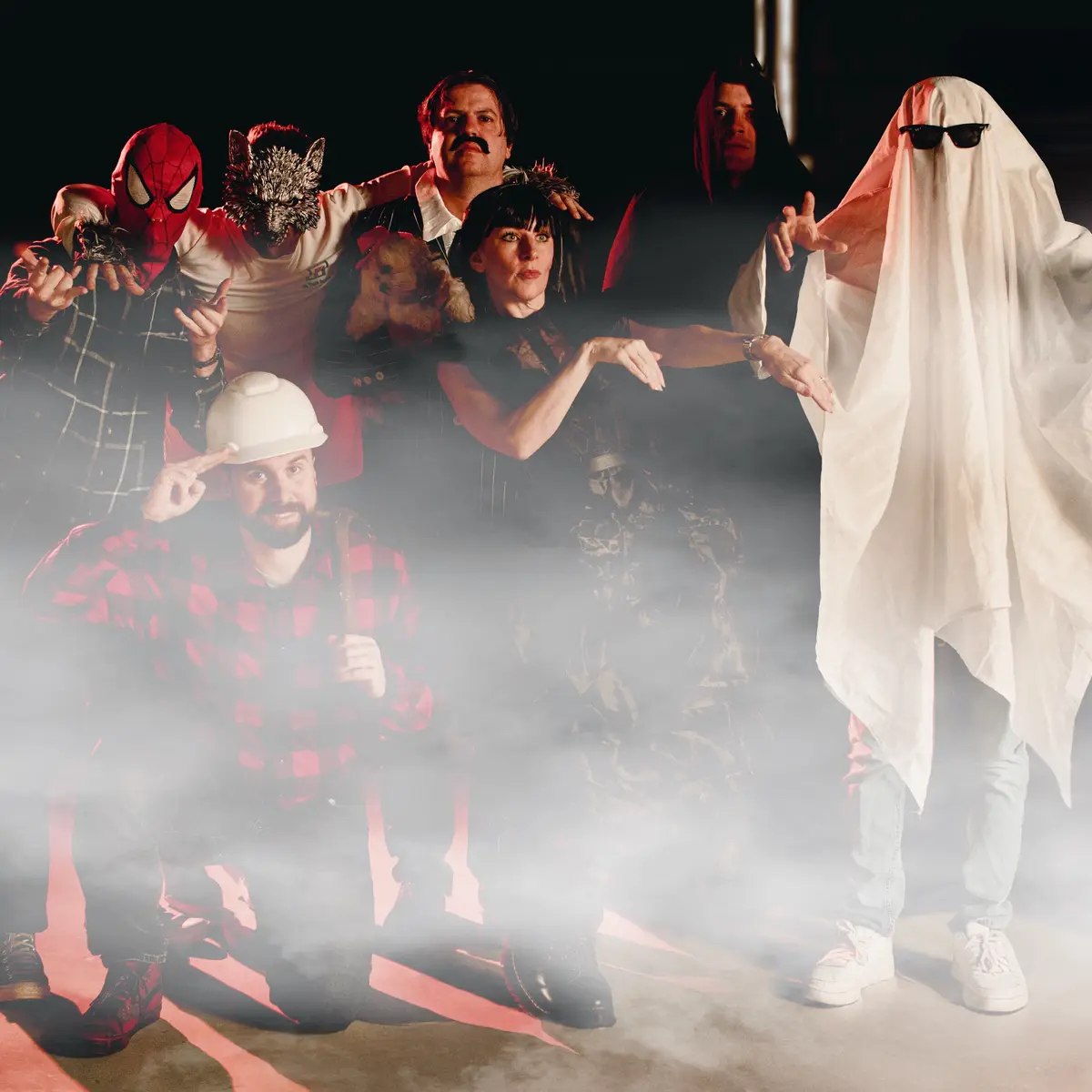 A group of seven people in various Halloween costumes pose together in a smoky, dimly lit setting. From left to right: a…