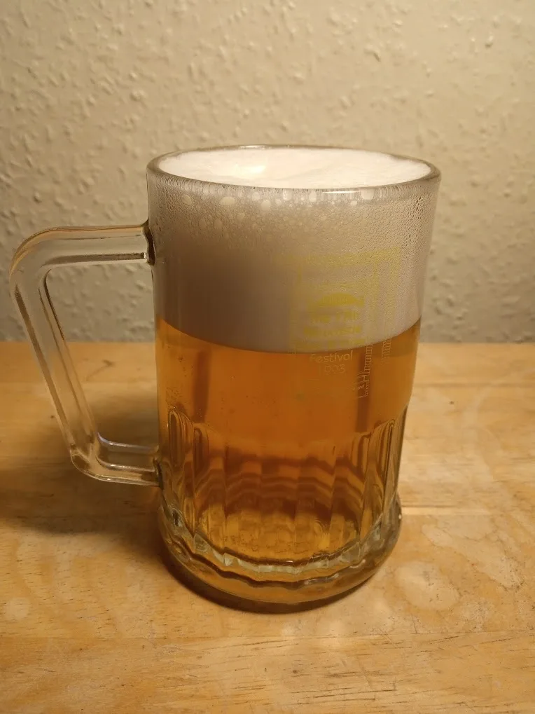 A clear glass beer mug with a handle sits on a wooden surface, filled with golden beer topped by a thick white foam head…