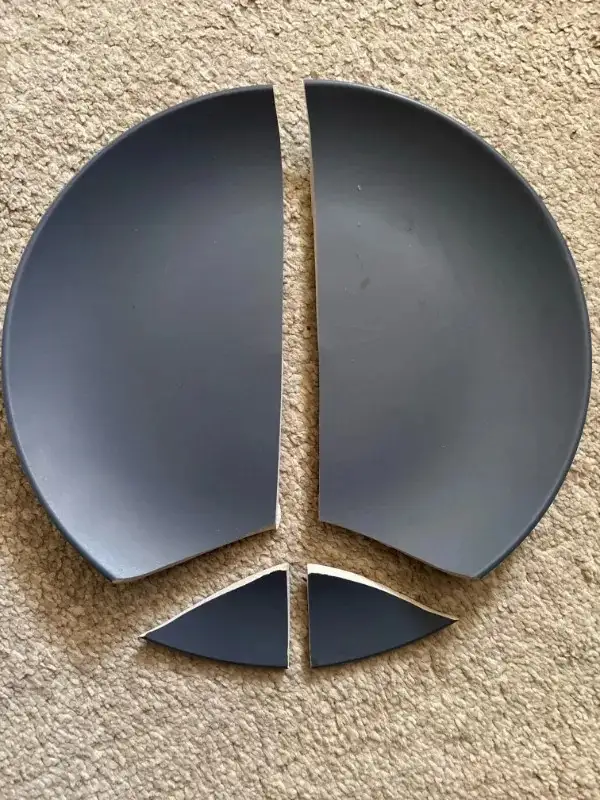 A dark gray plate is shattered into four pieces on a beige carpet, arranged to vaguely resemble a face with two large…