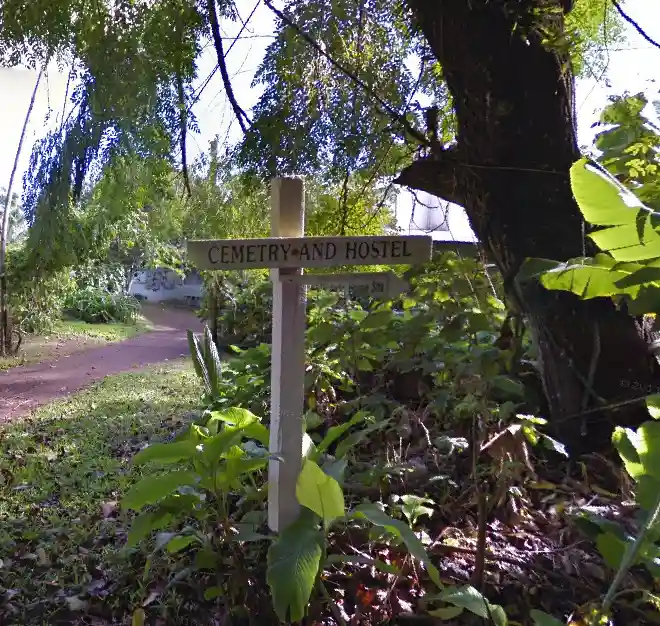 A wooden cross-shaped sign in a lush, green outdoor setting reads 'CEMETERY AND HOSTEL'. The sign is surrounded by foliage…