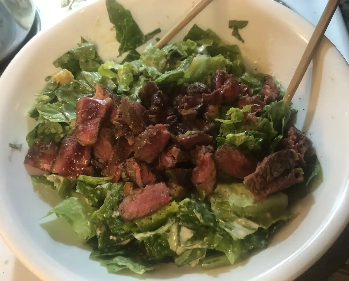 A close-up photo shows a white bowl filled with a salad of green lettuce leaves, croutons, and a creamy dressing, topped…
