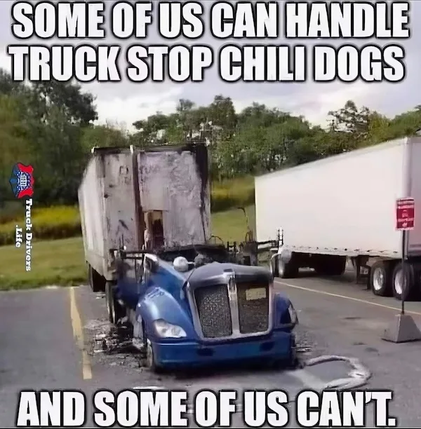 A blue semi-truck is severely damaged, with its front end crushed and melted, parked in a truck stop lot next to another…