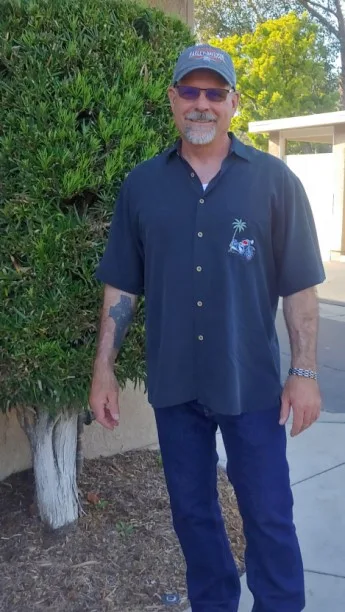 A middle-aged man with a goatee, sunglasses, and a baseball cap stands outdoors in front of a green bush. He wears a dark…