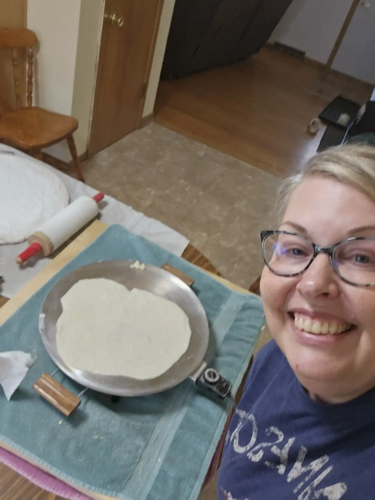 A smiling woman with glasses takes a selfie next to a tortilla press with a freshly made tortilla on it. A rolling pin and…