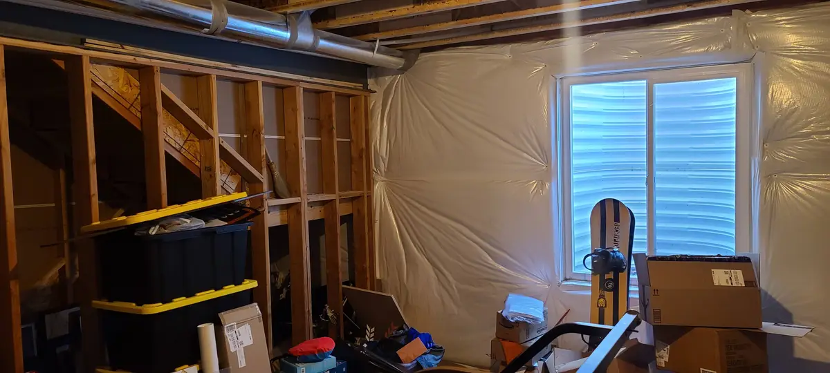 A messy, unfinished basement with exposed wooden framing, plastic sheeting covering walls, and various boxes and storage…