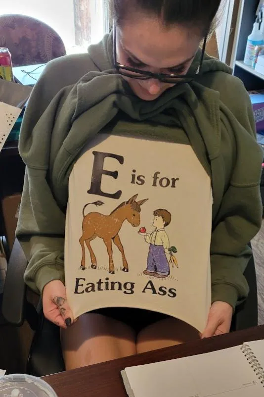 A person wearing a green hoodie holds up a beige t-shirt that reads 'E is for Eating Ass' with an illustration of a donkey…
