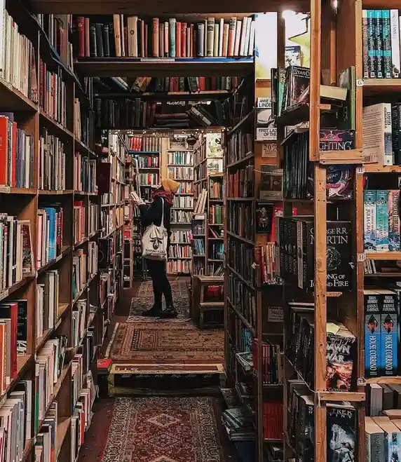 A person wearing a yellow beanie and carrying a white tote bag stands in the middle of a narrow, book-filled aisle in a…