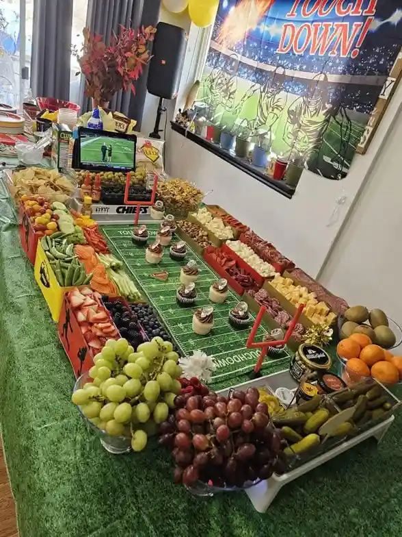 A football-themed party spread features a long table covered in green artificial turf, with food arranged like a football…