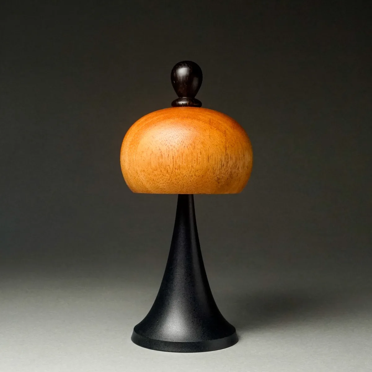 A minimalist wooden object with a light-colored, rounded dome top and a dark, conical base stands against a plain gray…