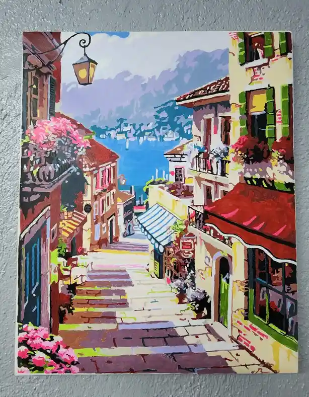 A colorful painting depicts a picturesque European street with stairs leading down to a waterfront, featuring buildings with…