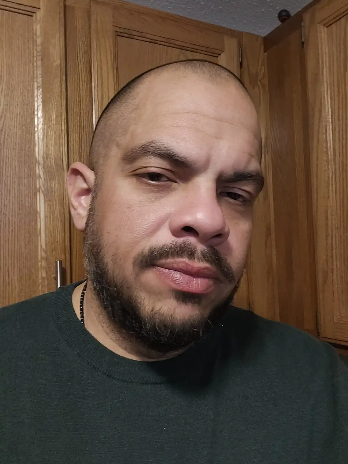 A man with a shaved head and goatee stares directly at the camera with a deadpan, unimpressed expression. He wears a dark…