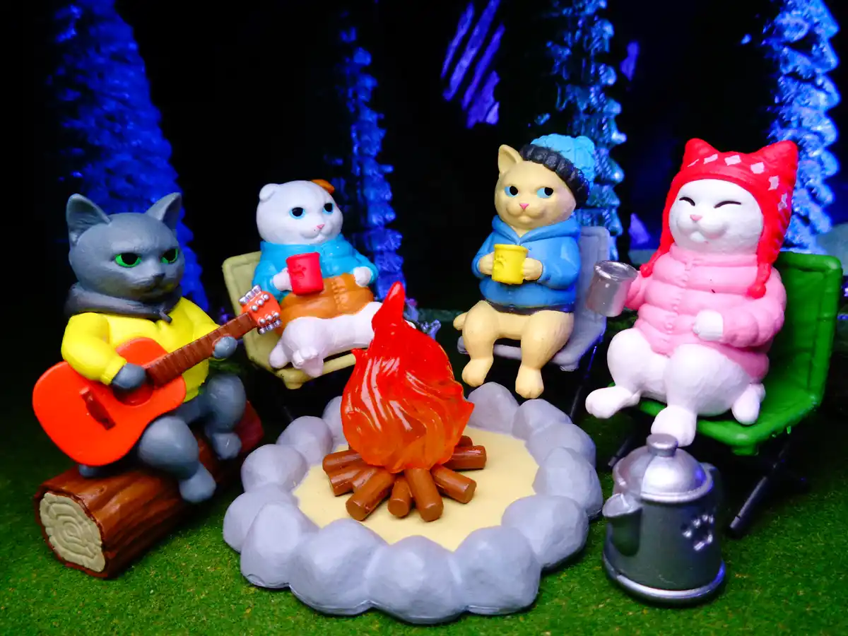 Four plastic cat figurines are gathered around a miniature campfire in a forest setting, each dressed in cozy winter clothes…