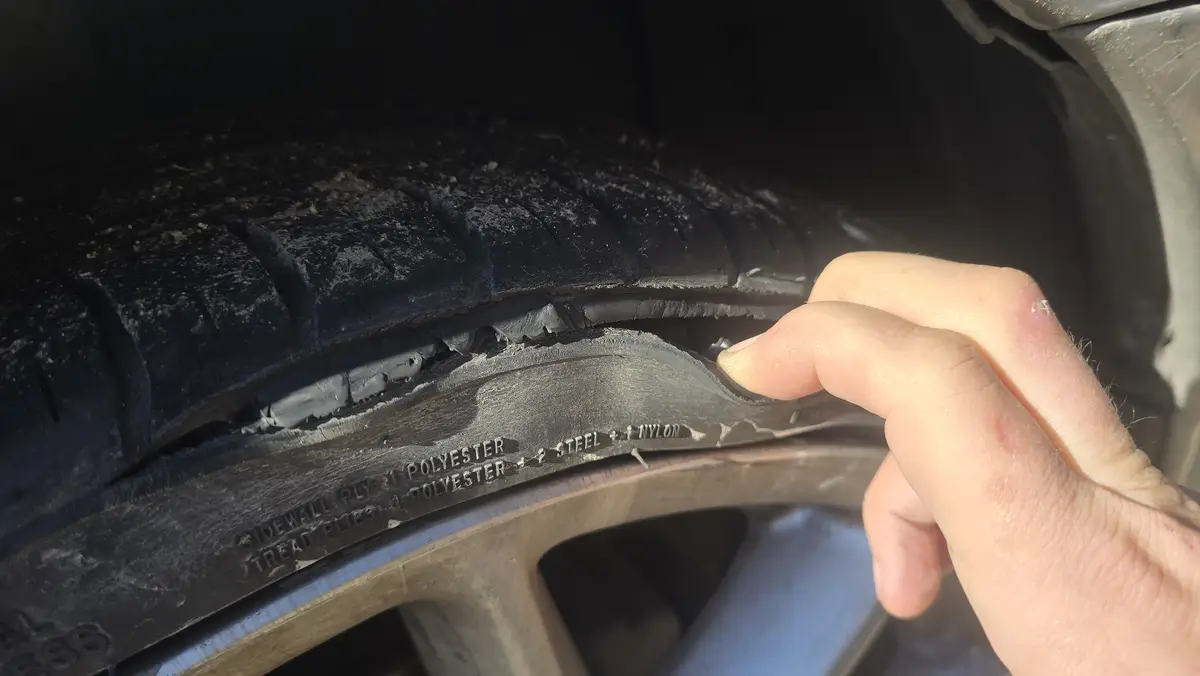A close-up photo shows a person's hand pointing to a damaged car tire, where the sidewall is visibly cracked and peeling…