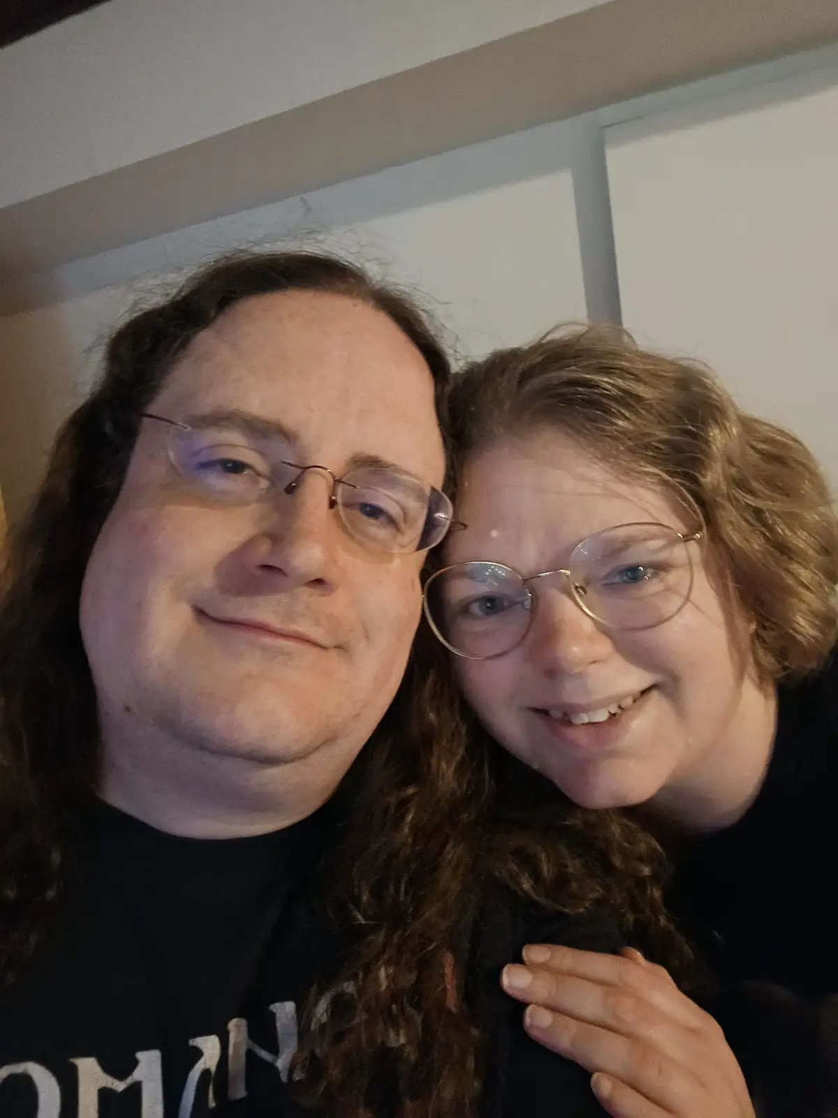 A selfie of two people, a man with long brown hair and glasses and a woman with short blonde hair and glasses, smiling…