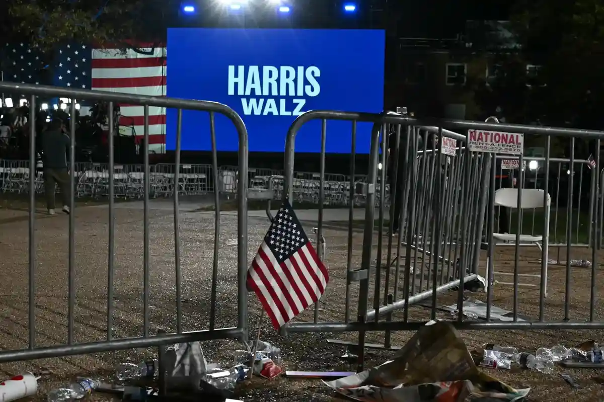 A nighttime photo of an outdoor political rally with a large blue screen displaying 'HARRIS WALZ' in white text. In the…