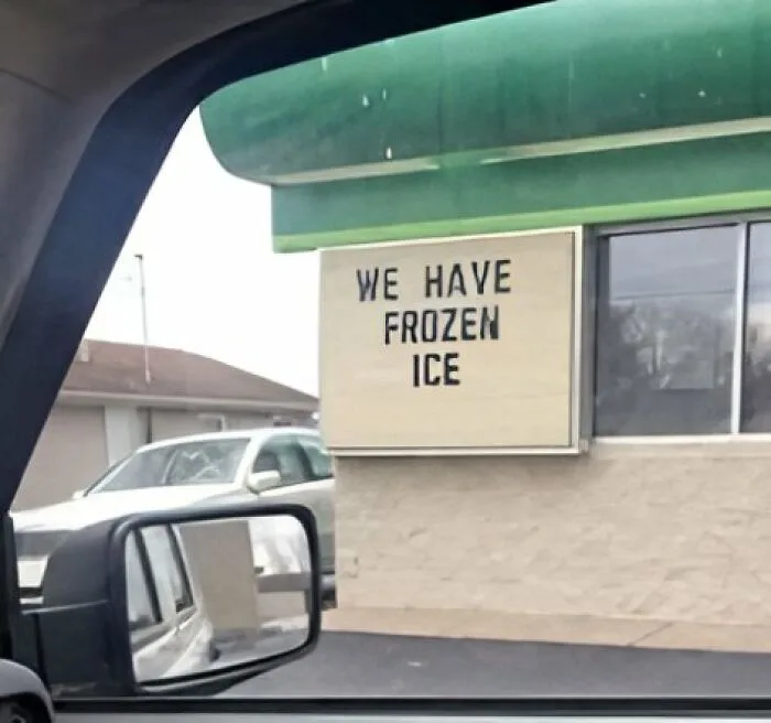 A photo taken from inside a car shows a beige sign on the exterior wall of a building with a green awning. The sign reads…