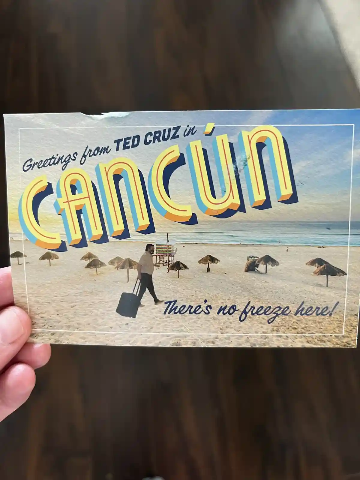 A hand holds a vintage-style postcard depicting a sunny beach scene in Cancún with thatched umbrellas and a man walking with…