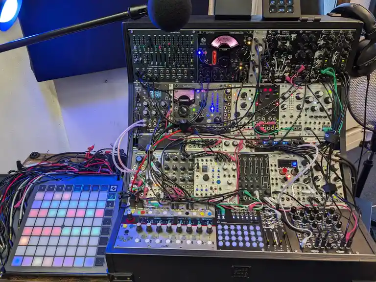 A densely packed modular synthesizer setup with numerous black and silver modules, glowing knobs, and a tangle of colorful…