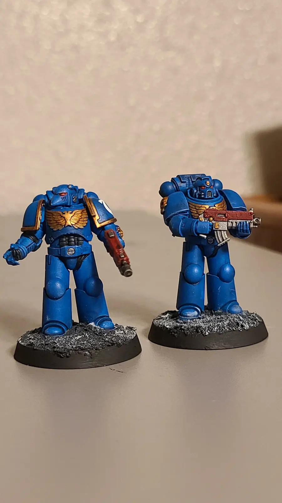 Two blue-painted Warhammer 40,000 Space Marine miniatures stand side-by-side on textured bases, one holding a weapon and the…