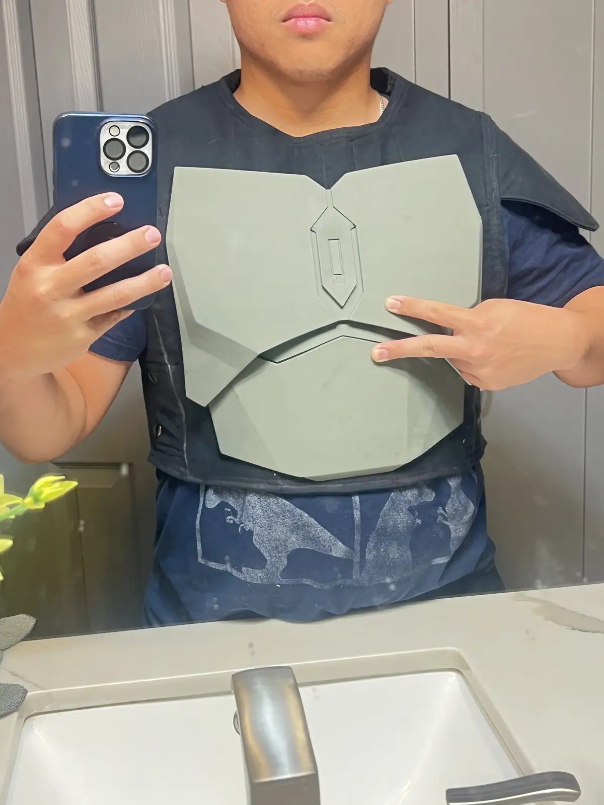 A person takes a mirror selfie in a bathroom while wearing a grey, angular, sci-fi style chest plate over a dark t-shirt…