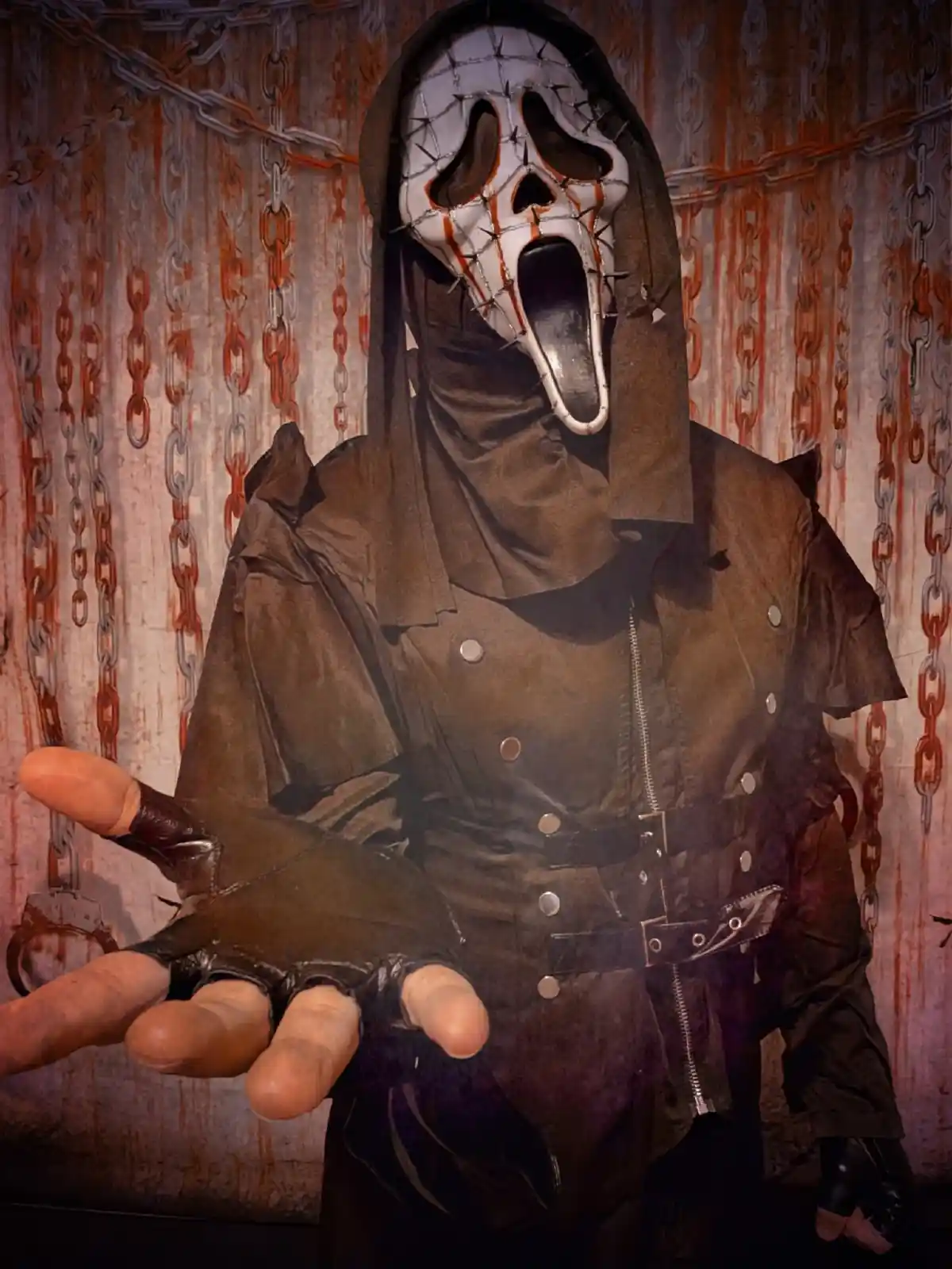 A person in a horror movie costume featuring a white, screaming ghost face mask with barbed wire and blood stains, wearing a…