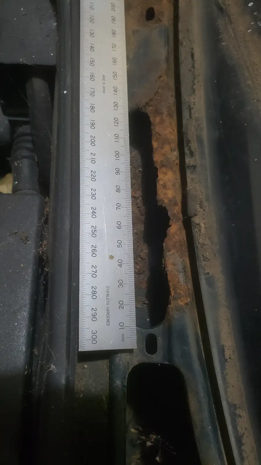 A close-up photo shows a metal ruler placed against a large, jagged hole in what appears to be a car's undercarriage or…