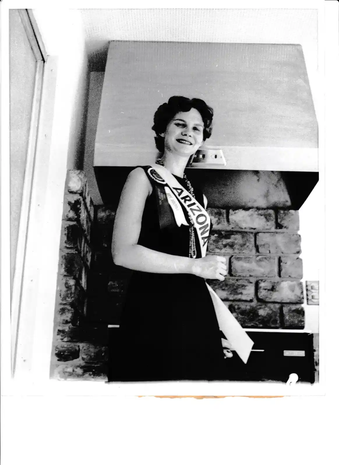 A black-and-white photo shows a smiling woman wearing an 'Arizona' sash, standing in a kitchen next to a brick wall and a…