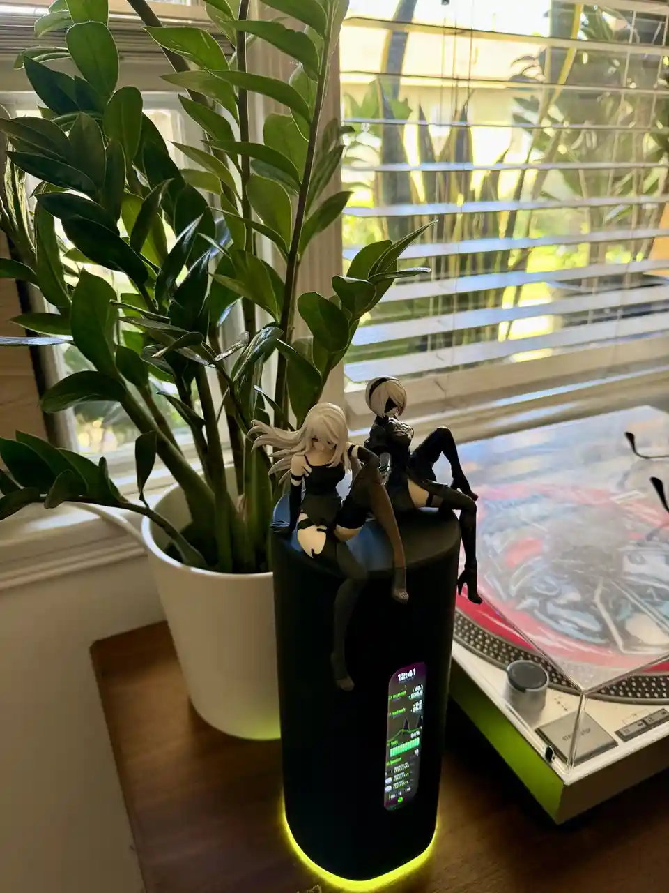 Two anime figurines, 2B and 9S from NieR:Automata, are posed sitting on top of a black smart speaker with a glowing yellow…