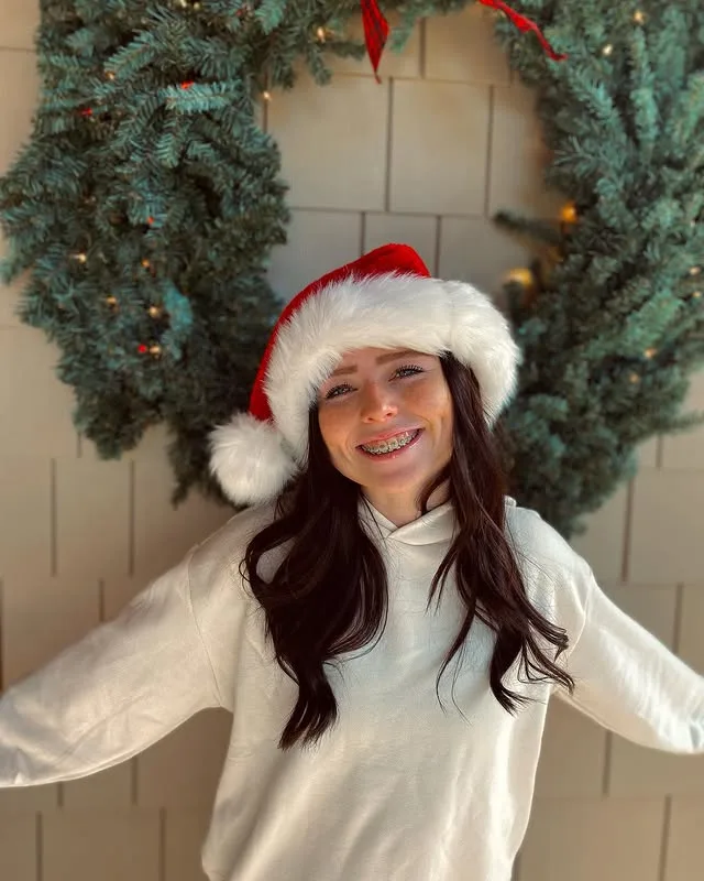 A young woman with dark hair and visible dental braces smiles cheerfully while wearing a red and white Santa hat. She is…