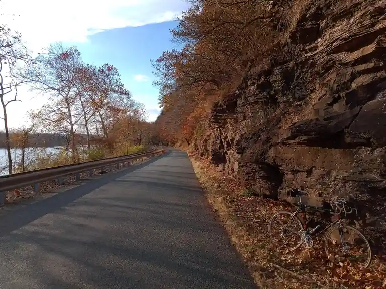 A black road bicycle leans against a rocky cliffside beside a paved path in autumn, surrounded by fallen leaves and trees…