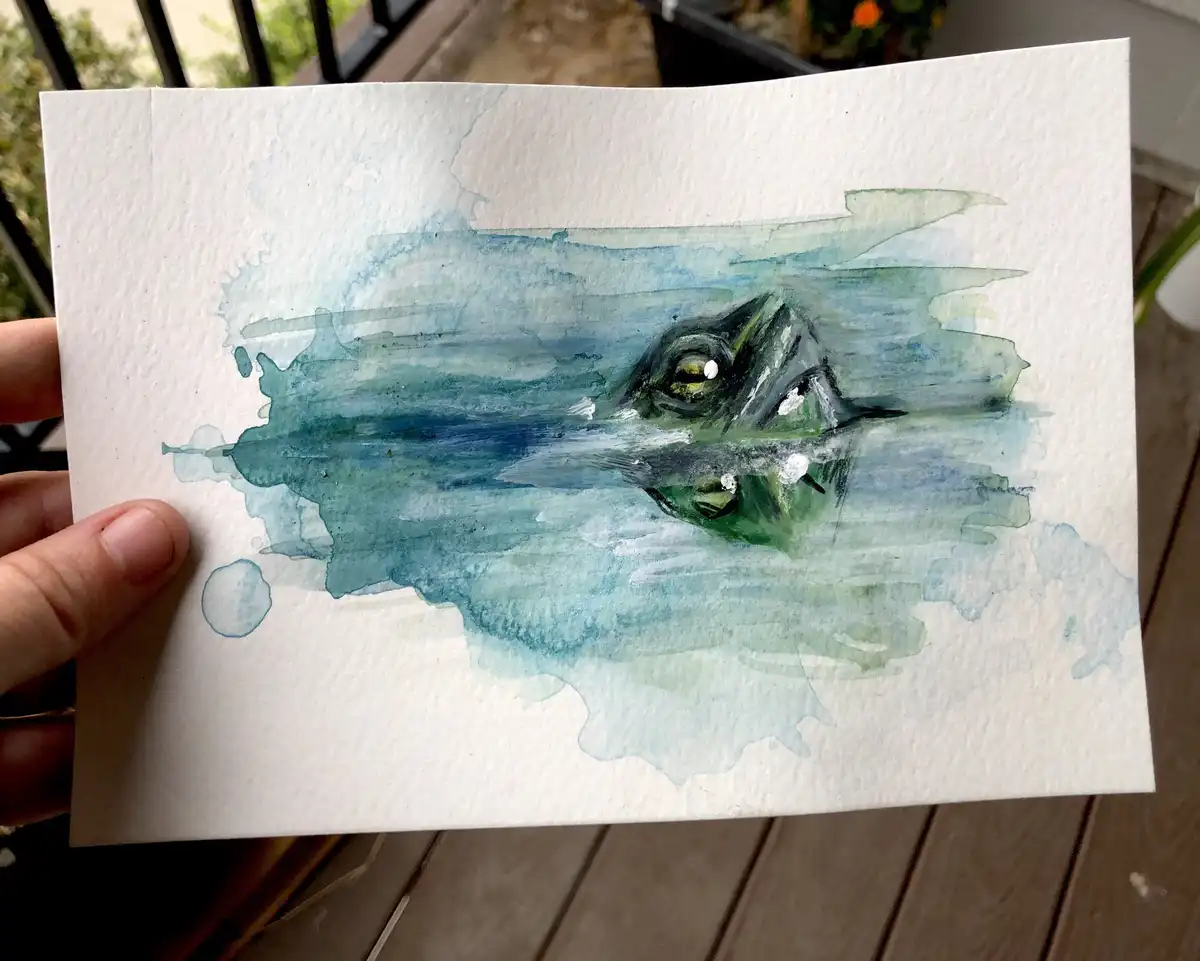 A hand holds a watercolor painting on textured white paper depicting a turtle's head emerging from blue-green water, with…